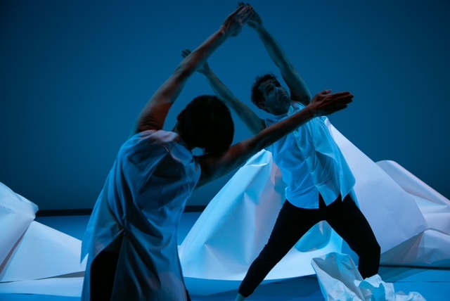 Two dancers in sleeveless white blouses tilt their bodies sideways and extend their arms up into a V amidst paper sculptures.